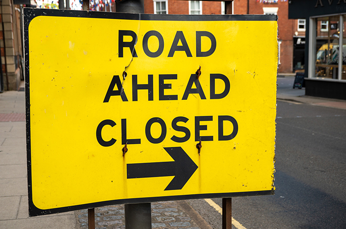 A bright yellow road sign reads ‘Road Ahead Closed’