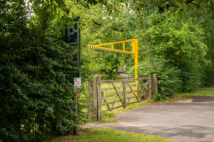 Closed five-bar gates at the entrance to the Fishpond Meadow car park where a Pay and Display sign is partially covered by leafy greenery