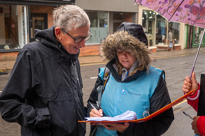 A woman with folder and pen in her hands stands next to a smiling grey haired man as she asks him survey questions