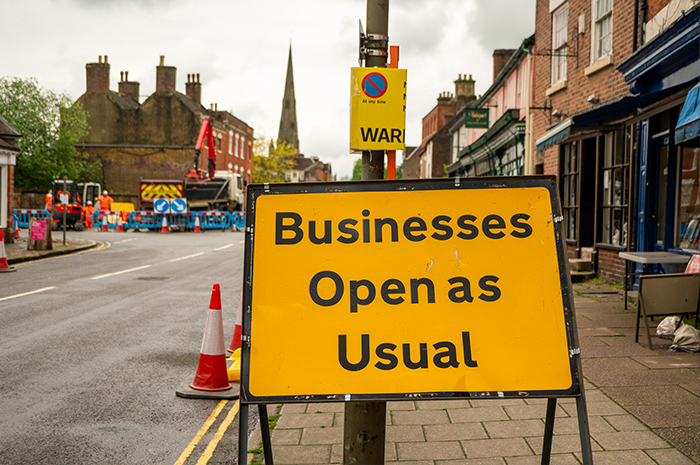 A large yellow road sign reads 'Businesses Open as Usual', traffic cones and road work barriers are visible in the background 